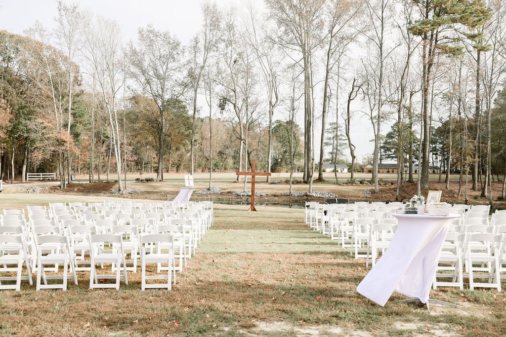 Outdoor wedding ceremony setup with white chairs arranged in rows on grass, a wooden cross at the front, and tables with tablecloths and decorations on either side, surrounded by trees.
