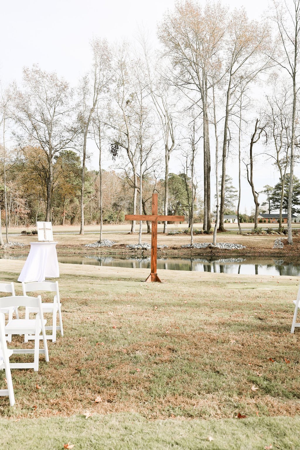 An outdoor wedding setup with white chairs, a wooden cross, and a small table with a white cloth near a pond