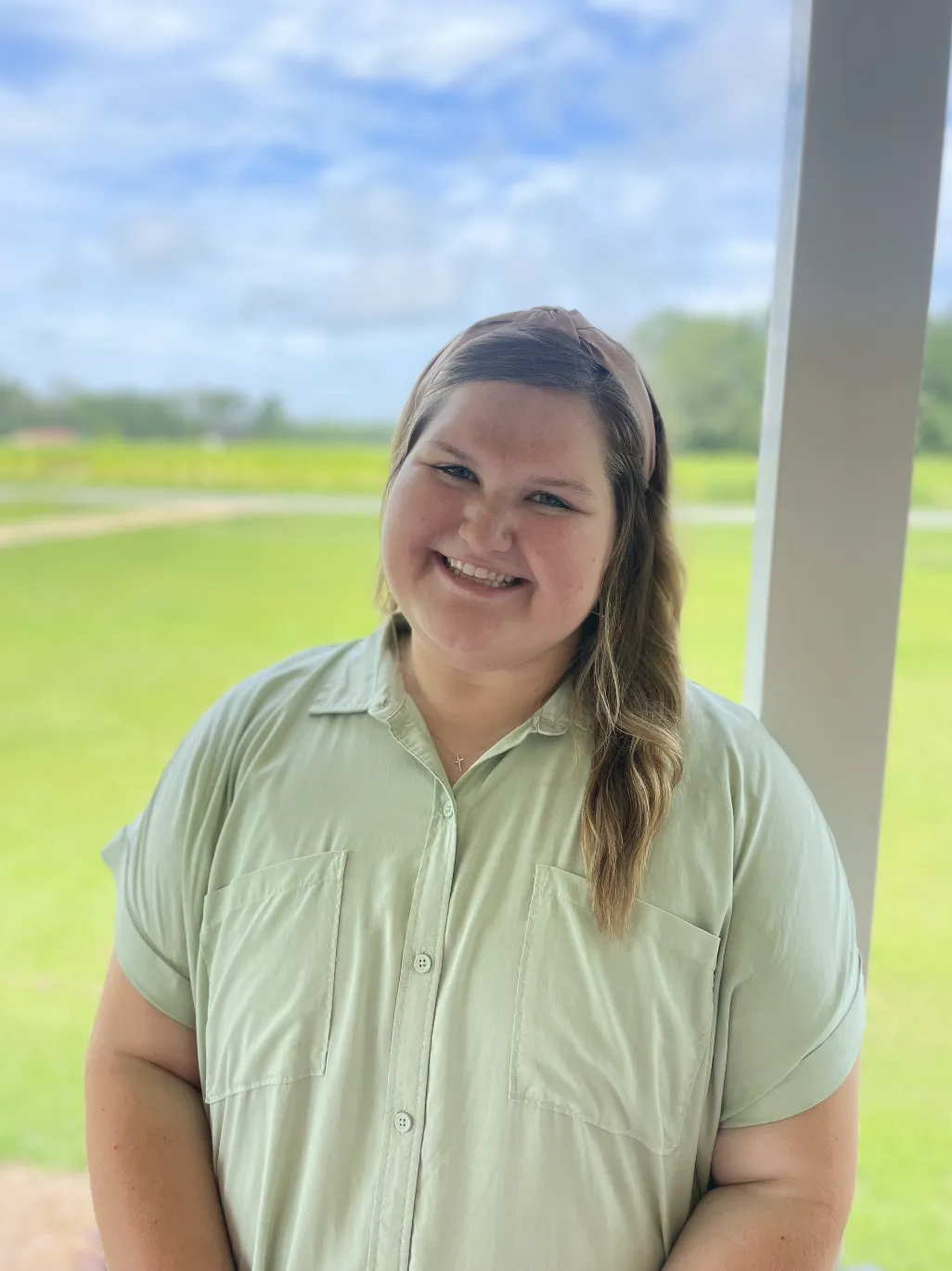 A smiling girl with light skin and long wavy hair, wearing a beige headband and a light green button-up shirt, standing outside with green fields and blue sky with clouds in the background.