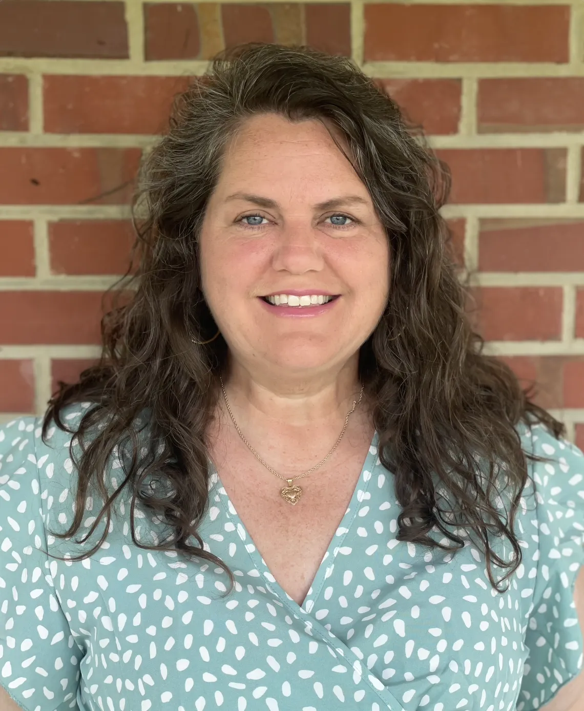 A woman with long wavy brown hair and blue eyes smiling in front of a brick wall, wearing a teal top with white polka dots and a gold heart necklace.