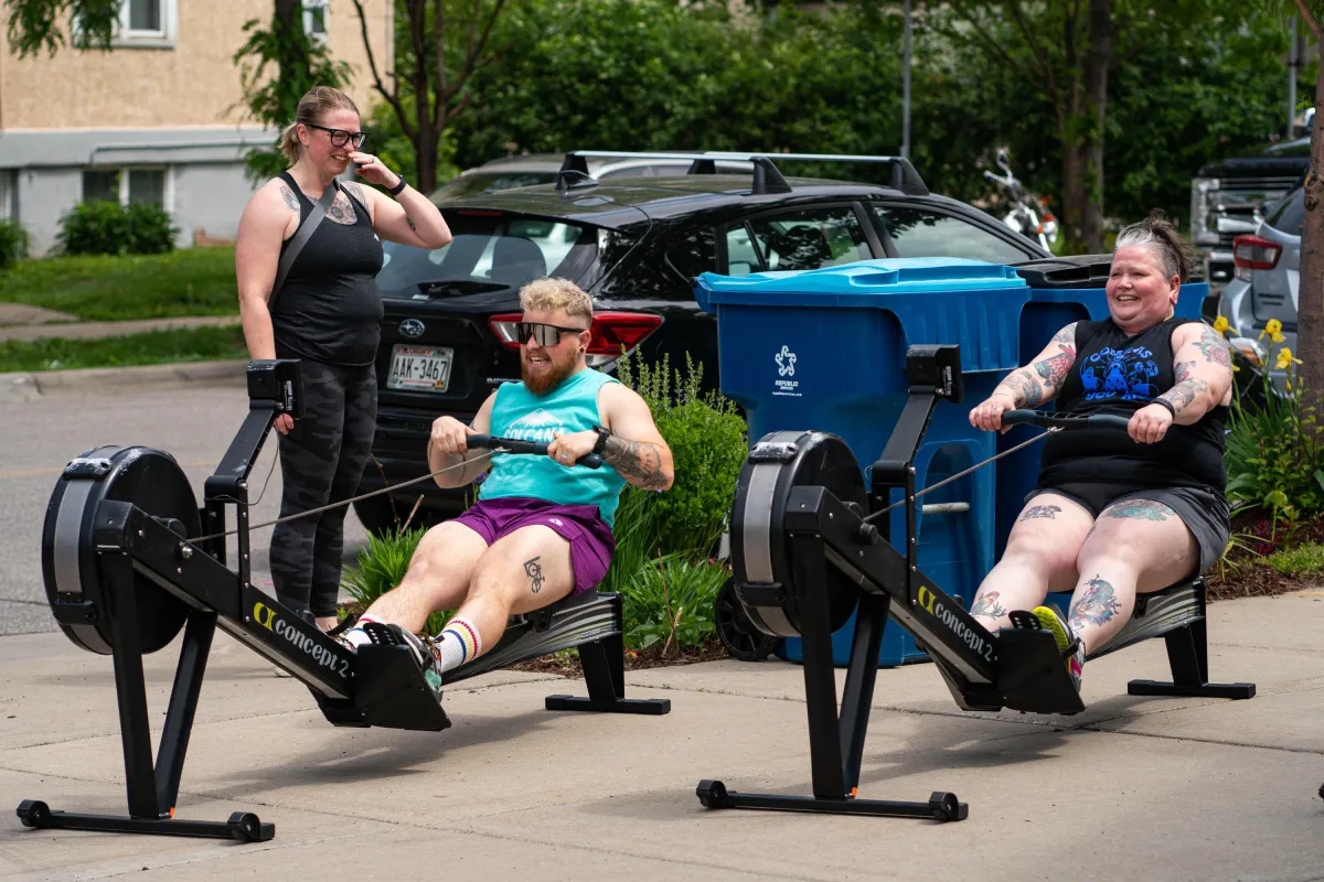 Two people rowing on rowing machines out in the sun in front of a food truck and garden.