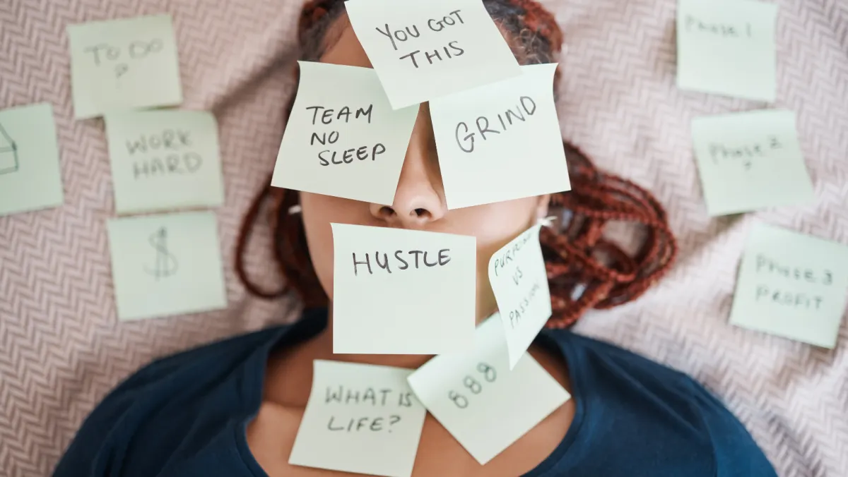 The image shows a person lying down with multiple sticky notes placed on their face and surrounding area. The notes have motivational and work-related messages written on them, such as "You got this," "Grind," "Hustle," "Team No Sleep," "Work Hard," "What is life?," "To do," "$," "Phase 1," "Phase 2," "Phase 3 = Profit," and "Purpose vs. Passion." The person appears to be overwhelmed or exhausted.