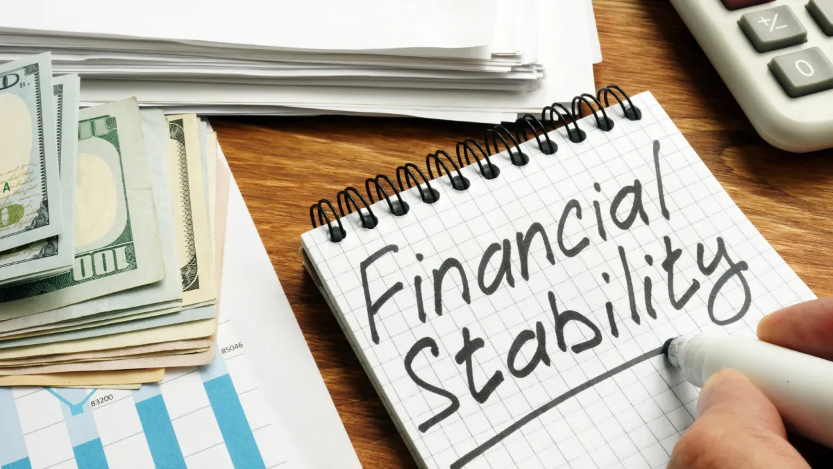 A close-up image showing a hand writing "Financial Stability" in a notebook with a marker. Nearby are stacks of US dollar bills, financial documents, and a calculator, all placed on a wooden table. The image emphasizes the concepts of financial planning, budgeting, and achieving economic security. Ideal for blog posts on financial tips, stability strategies, and money management.