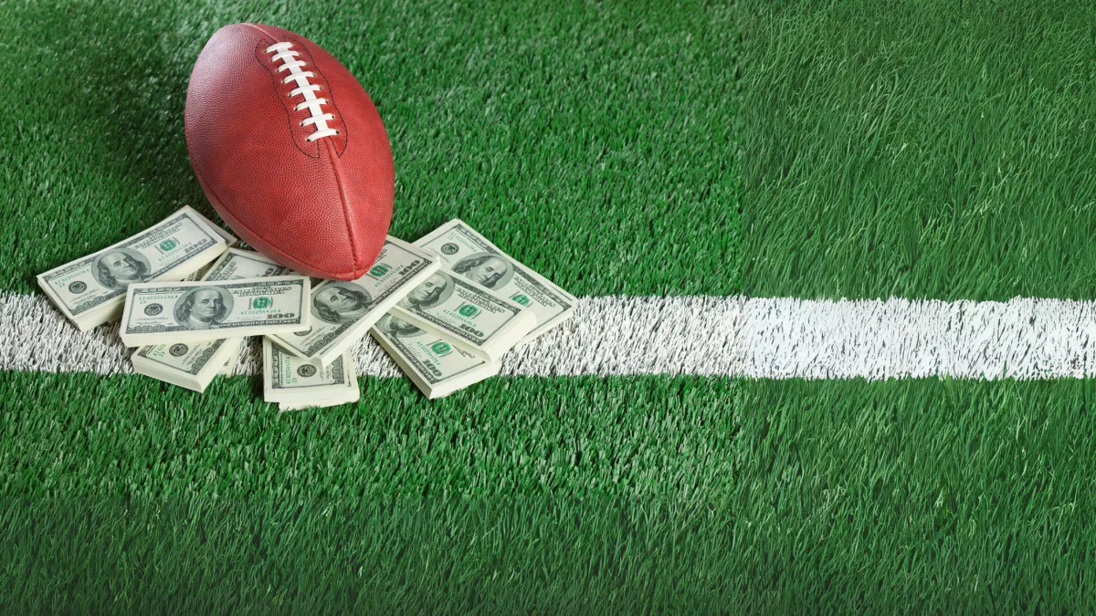 A football rests on a pile of U.S. dollar bills on a grassy field near a white line.