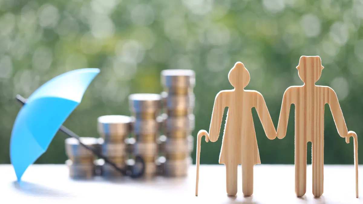 Wooden cut-outs of an elderly couple holding hands stand in front of stacked coins and a blue umbrella against a blurred green background. The figures are symbolic, representing retirement planning, with the woman holding a walking stick