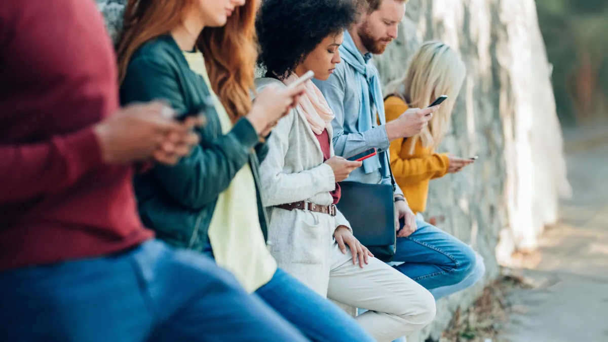 A group of five people sits outdoors, leaning against a stone wall, all looking at their smartphones.