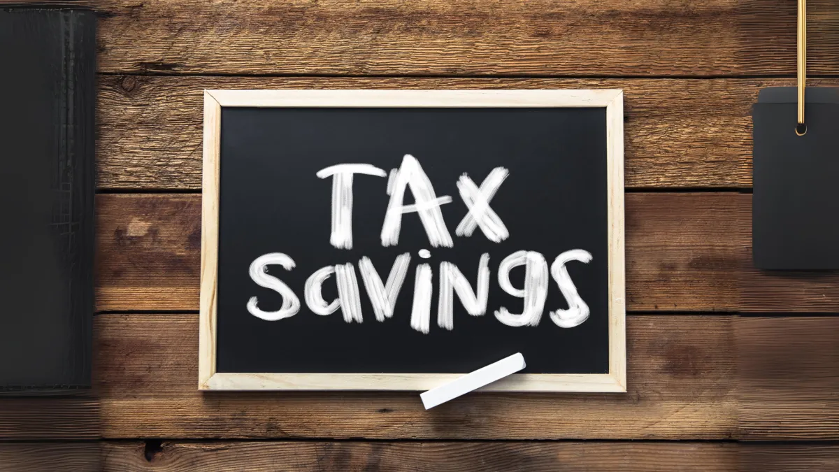 A small chalkboard with a wooden frame rests on a wooden surface. The board has "TAX SAVINGS" written on it in white chalk, with a piece of chalk placed on the bottom frame ledge. A black notepad and a price tag are partially visible on either side.s owner holding a compass next to a tax changes sign