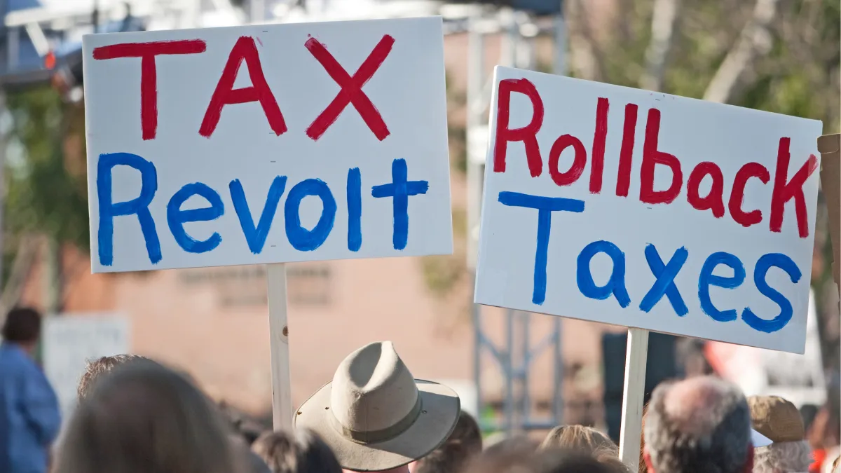 A crowd of people holding signs that read "TAX Revolt" and "Rollback Taxes" at a protest or rally.