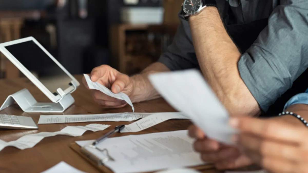 Two people review receipts and documents at a desk with a computer and a clipboard.