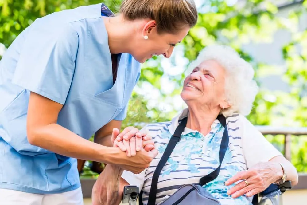 A smiling nurse in blue scrubs warmly holds hands with an elderly woman in a wheelchair, sharing a moment of kindness and connection in an outdoor setting.