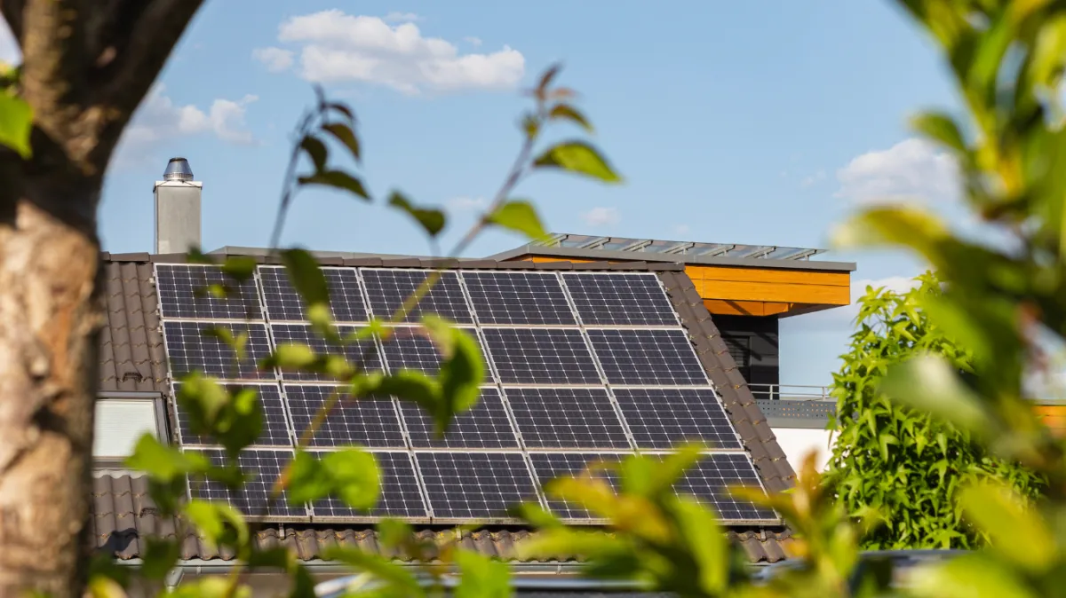 Solar panels installed on a tiled roof of a modern home, viewed through green foliage on a sunny day with blue sky and scattered clouds in the background.