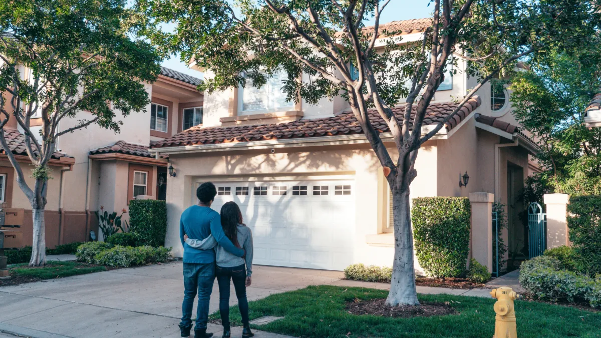 Young couple standing together looking at modern suburban home with tile roof and mature trees, representing homeowners considering gutter cleaning and maintenance decisions in Mildura