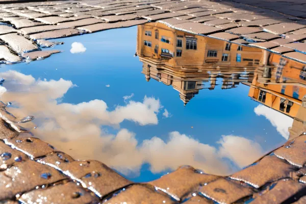 A close-up photograph of a clear rainwater puddle reflecting blue sky and clouds. The puddle is formed on a business district sidewalk or pavement, with the blurred outline of office buildings visible in the background