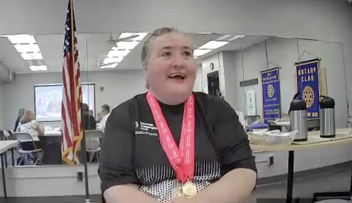 Amy Foster, wearing her Special Olympics Athlete Leadership shirt and several gold medals, is seated at the head table, engaged in conversation with present Riverbend Rotarians and those attending via Zoom.
