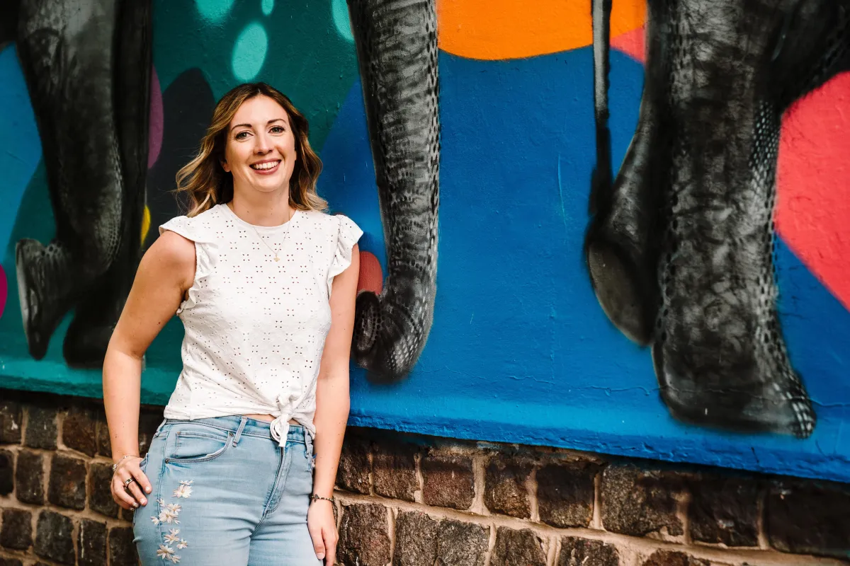 A brown-haired woman named Stephanie standing in front of a mural of what is presumably elephants.