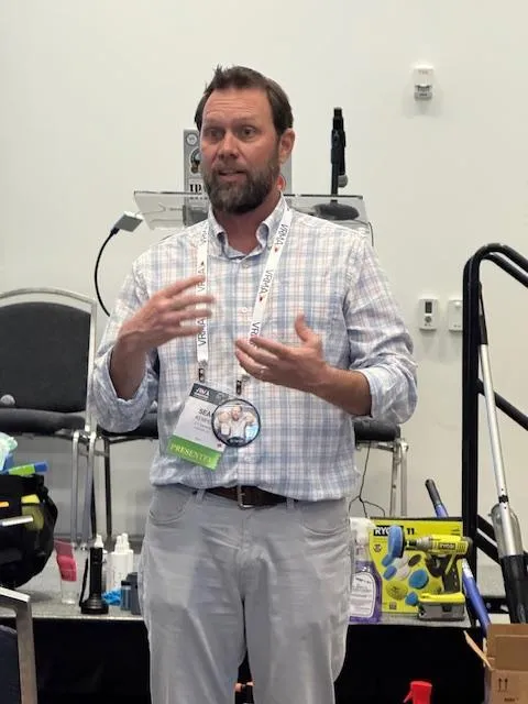 Sean Kemper stands at a podium, presenting "The Art of Cleaning" to an audience. He gestures toward a projected slide displaying cleaning chemistry concepts, with a table of cleaning products and pH test strips in the foreground. The setting is a well-lit conference room with attentive attendees.
