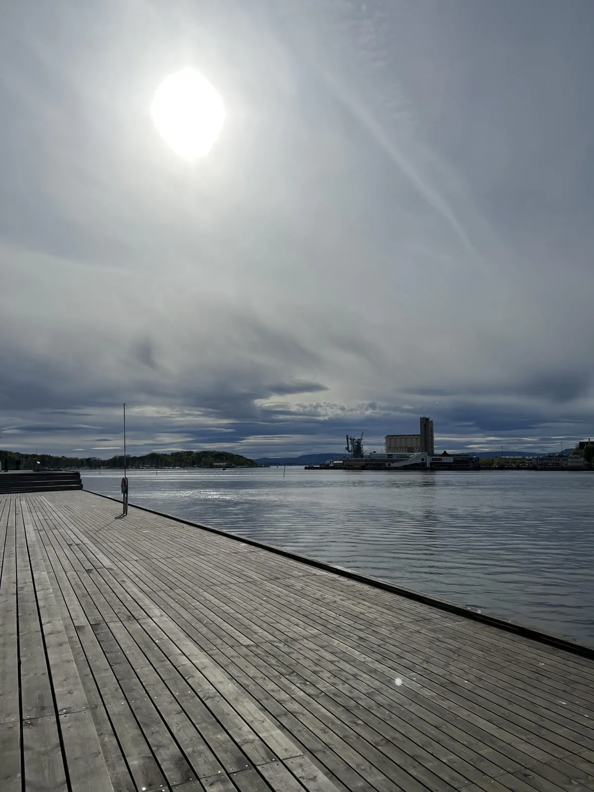 A boardwalk by a fjord with blue sky, clouds, and sunlight shining through.