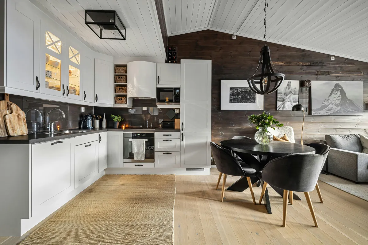 Stylish open-concept kitchen and dining area with a modern rustic design. The kitchen features white cabinetry with black handles, open shelving with wicker baskets, and built-in appliances. Dark wood paneling on the walls contrasts with a white shiplap ceiling, while warm lighting and decorative cutting boards add a cozy touch. A round black dining table with matching black velvet chairs and wooden legs sits beneath a contemporary pendant light. The adjacent seating area includes a gray armchair and monochrome mountain-themed wall art, creating a harmonious and inviting atmosphere.