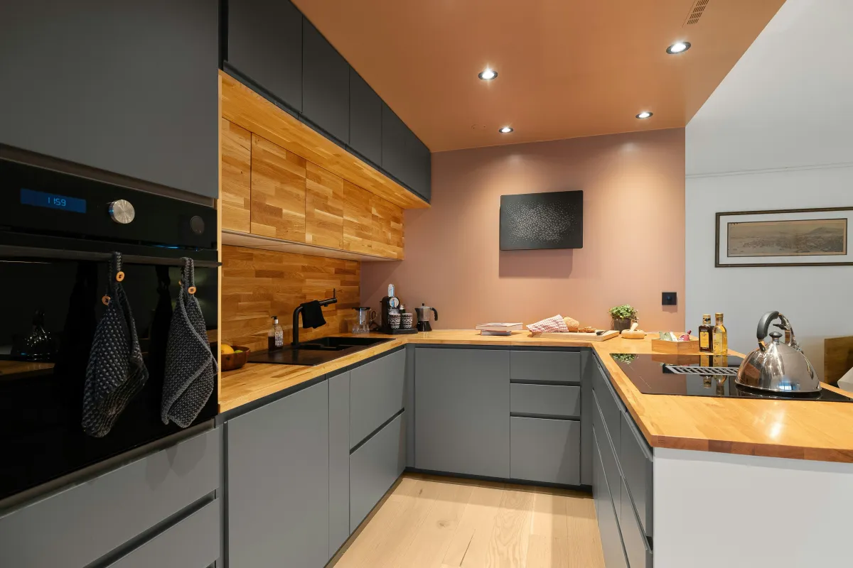 Modern modular kitchen featuring sleek matte grey cabinetry with minimalist handles, warm wooden countertops, and matching wooden backsplash. The kitchen is designed in a U-shape layout for efficiency and style. Built-in appliances, under-cabinet lighting, and soft recessed ceiling lights enhance the functionality and ambiance. A few kitchen essentials like a kettle, oil bottles, and a chopping board are neatly arranged on the counter, reflecting both practicality and aesthetic appeal.