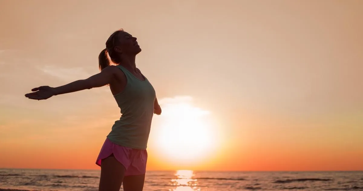 Woman standing with eyes closed and arms outstretched, with a sunset and ocean behind her, embodying transformation and renewal.