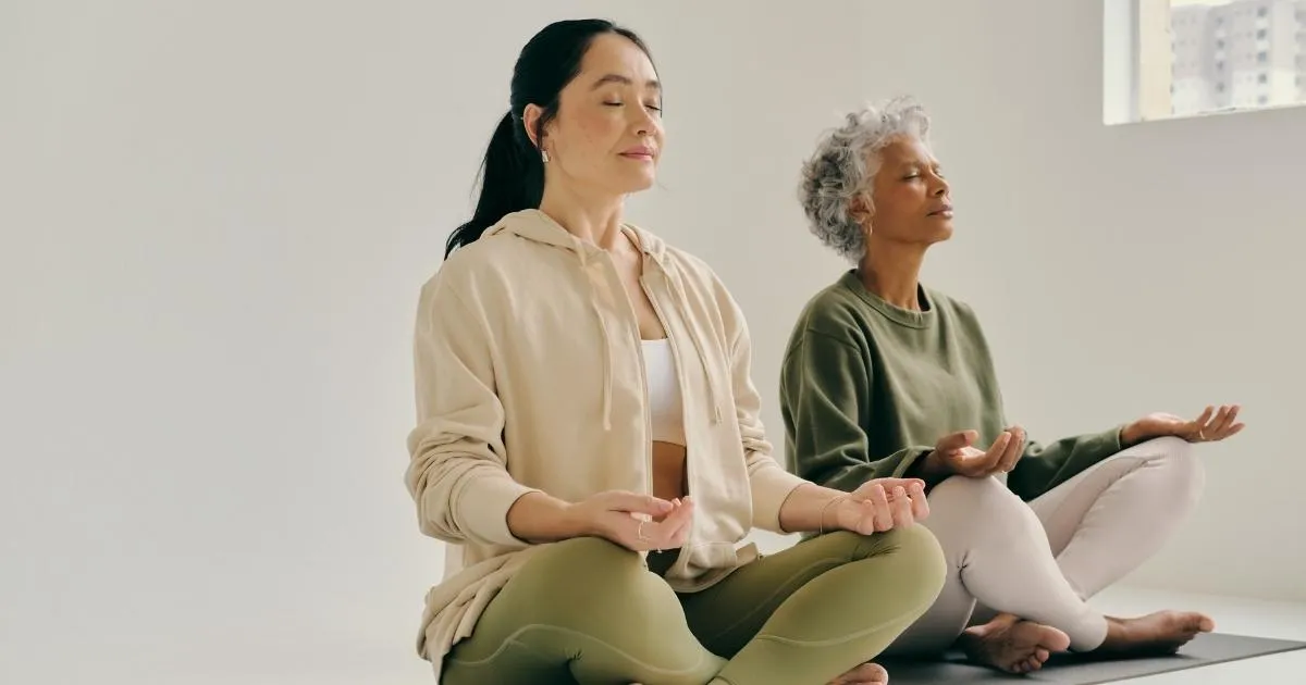 Two women sitting cross-legged on yoga mats in meditation, focusing on mindful movements and breath patterns to amplify core potential.