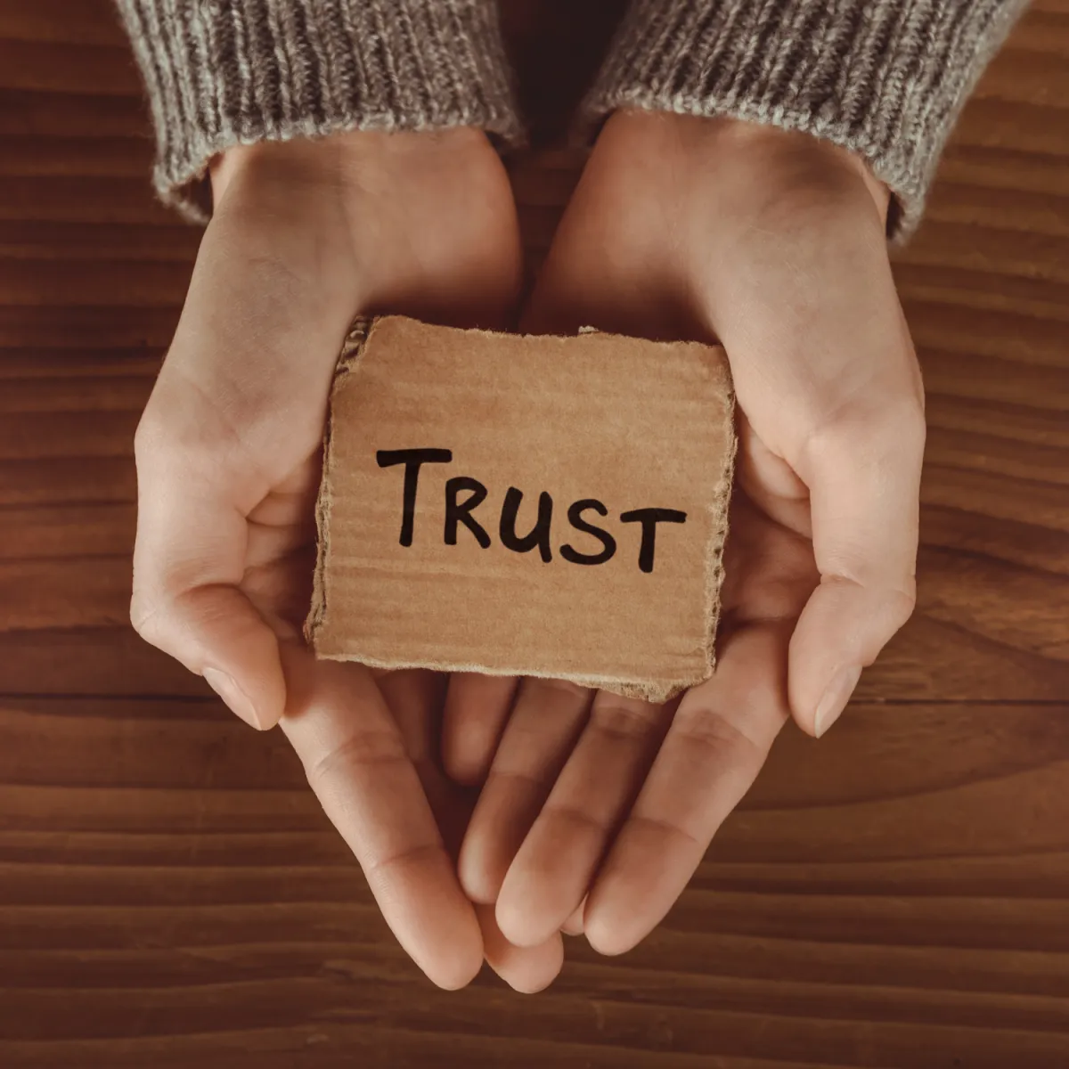 Two hands gently hold a piece of cardboard with the word "Trust" written on it. The hands are positioned against a wooden background, and the person is wearing a gray, knitted sweater.
