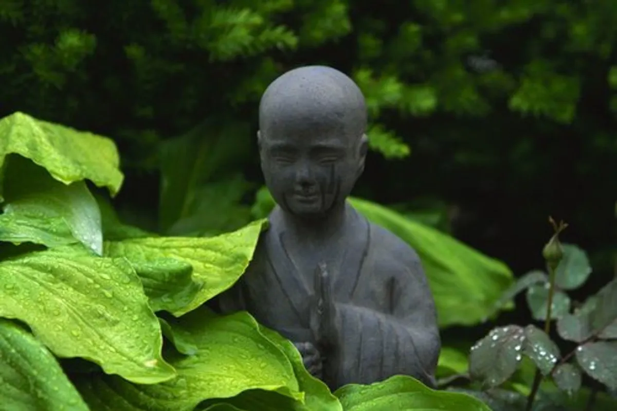 Dark stone Buddha figure with praying hands in amongst green leaves. A rain drop is on the face with the appearance of a tear