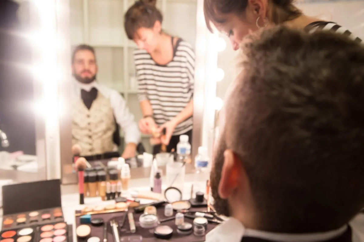 Mirror reflection of a man in light waistcoat and a makeup artist. There are stage lights around the mirror and thie image is slightly blurred