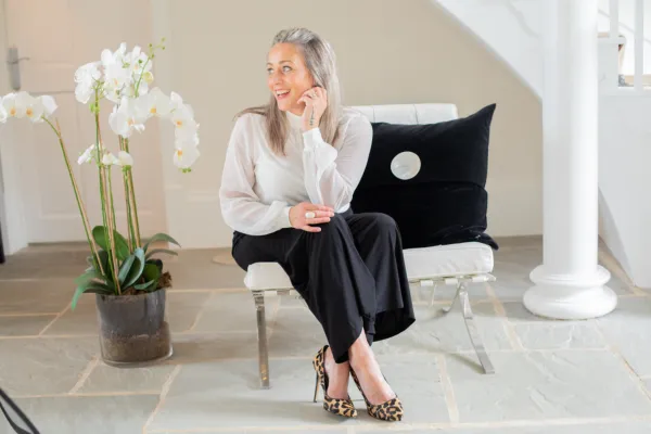 Lee-ann Cordingley sitting relaxed yet professional in a bright, elegant room. Wearing a white blouse, black trousers and leopard heels, she smiles to the side, blending yoga-teacher warmth with business-coach confidence. A white orchid sits beside her.