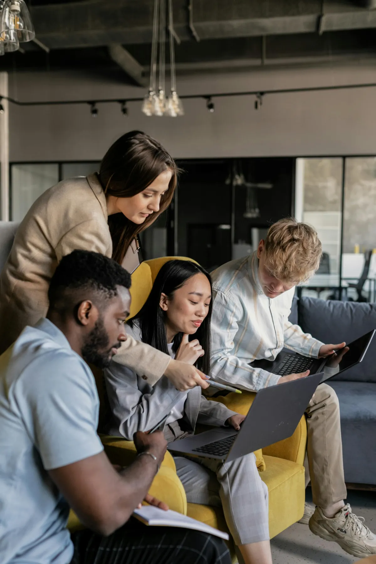 A diverse group of four young professionals collaborating in a modern office setting. They are sitting together, focused on their laptops, discussing and pointing at the screens, indicating a productive and engaging work environment.