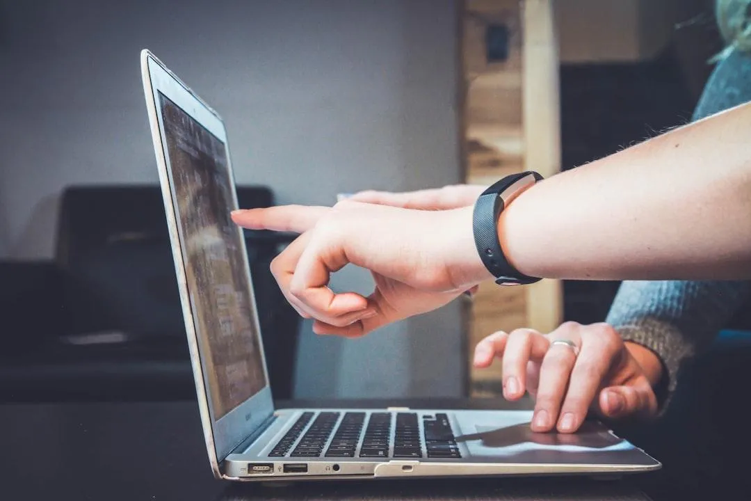 Close-up of two hands interacting with a laptop. One hand is pointing at the screen while the other hand is resting on the keyboard, indicating collaboration or instruction. The background is slightly blurred, creating focus on the hands and the laptop.