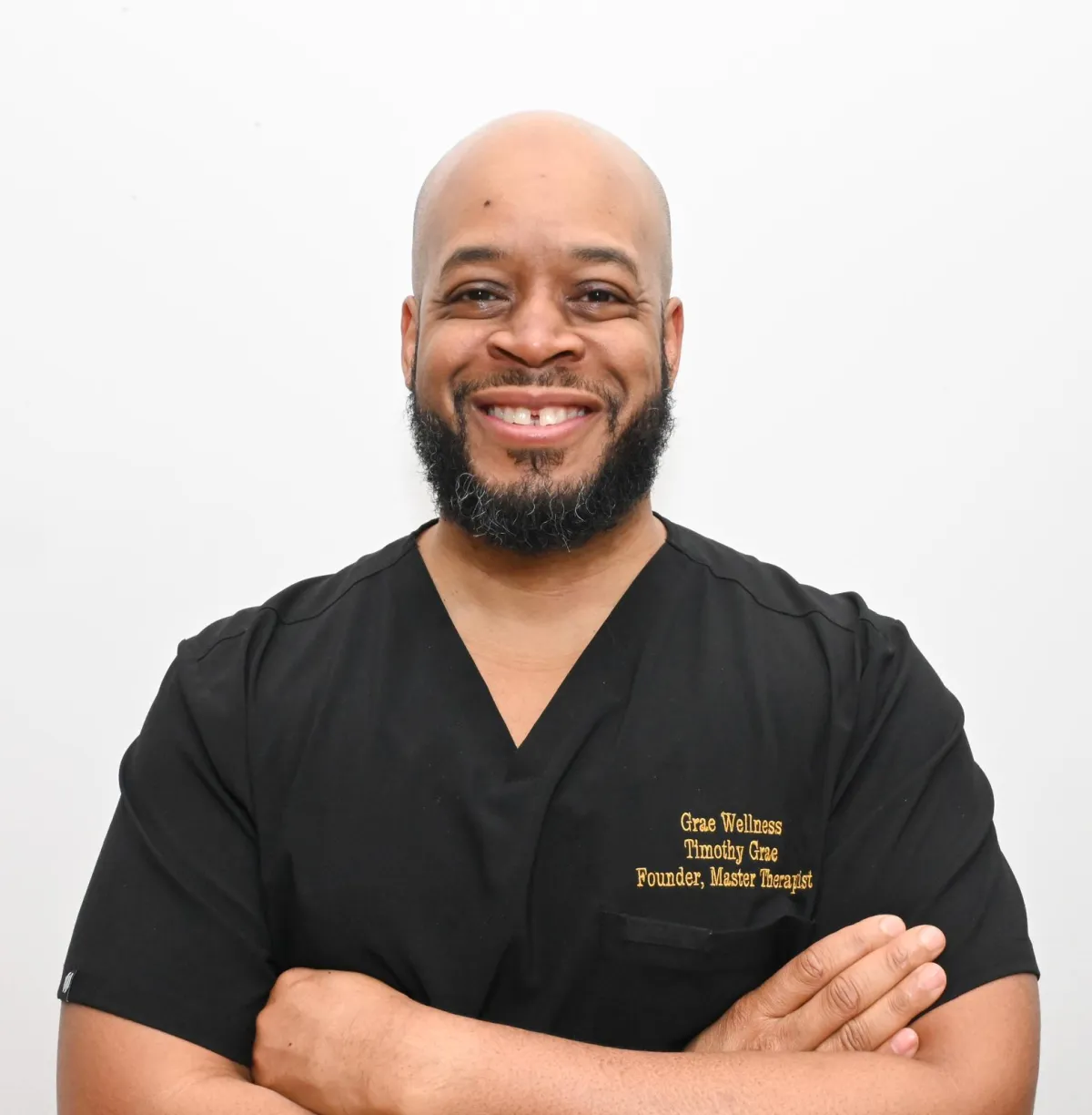 Professional headshot of Tim Grae, Founder and Master Therapist of Grae Wellness, smiling confidently in black scrubs with arms crossed against a plain white background.