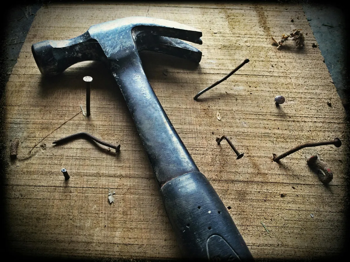 A close-up of a hammer resting on a wooden surface surrounded by bent and rusty nails, symbolizing repair and hard work.