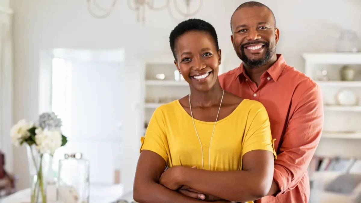 Picture of a black couple with the husband hugging from behind and they both smile