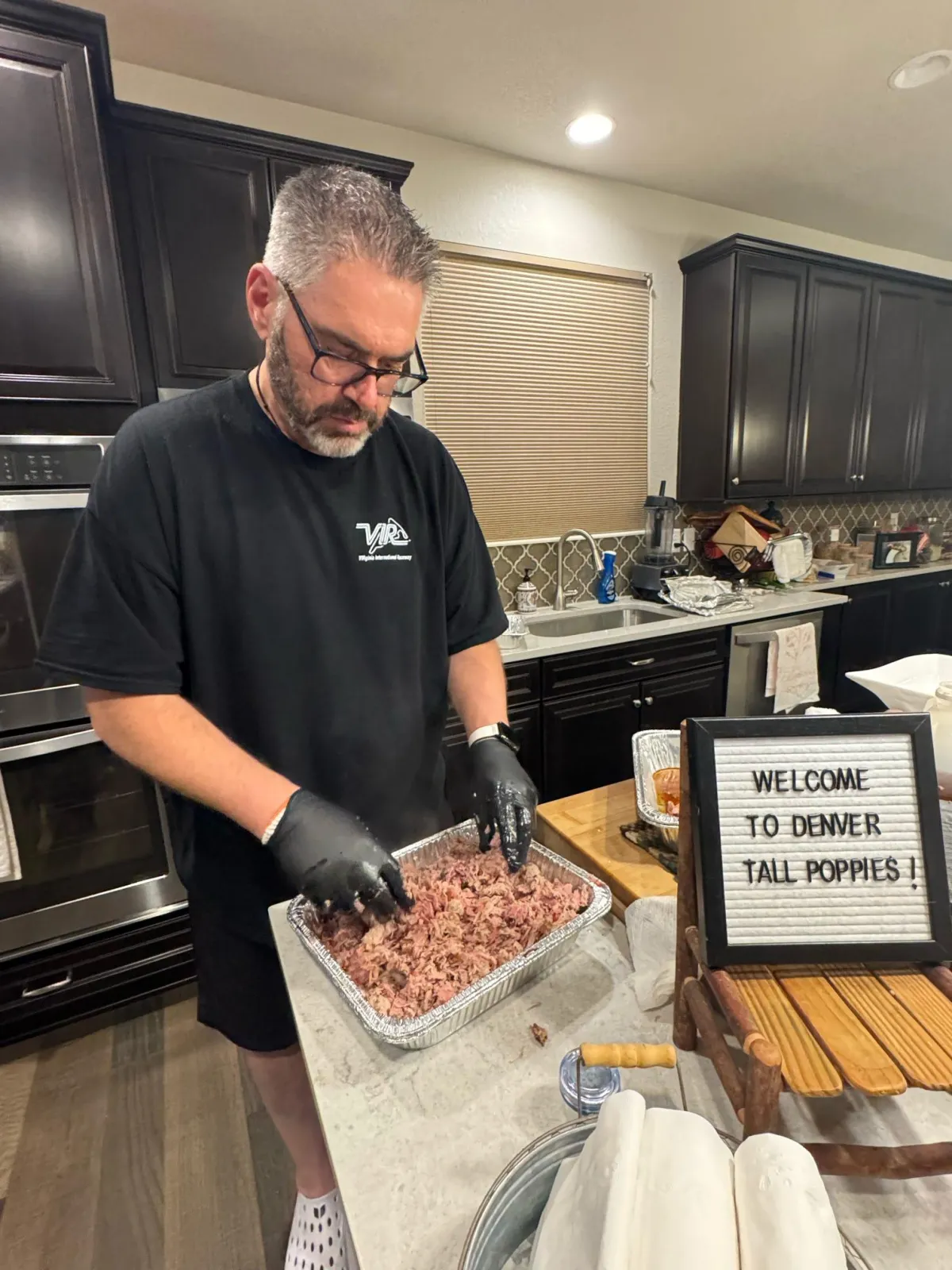 Man wearing gloves preparing seasoned meat in a kitchen, standing beside a sign that reads “Welcome to Desert Fox Popups.”