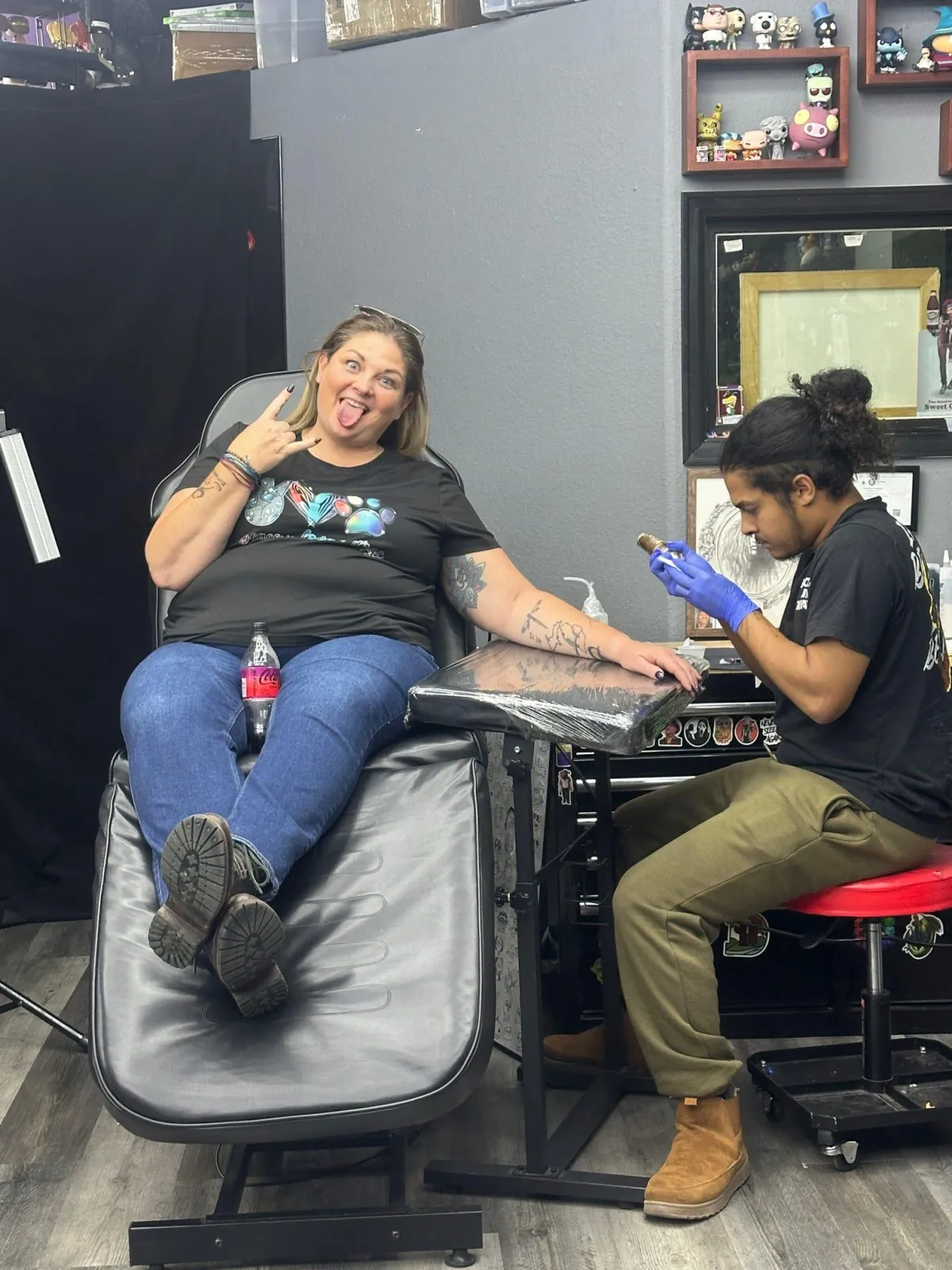 Woman lying on a tattoo chair while a tattoo artist works on her arm in a studio setting.