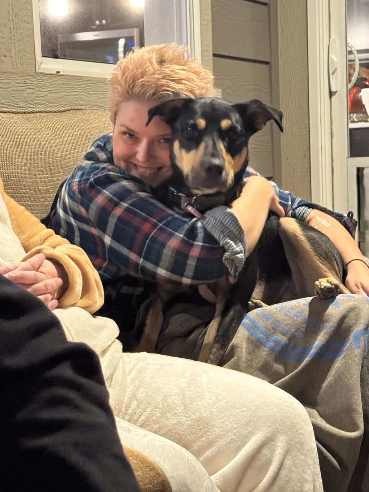 Person cuddling a black and tan dog on a couch in a cozy living room setting.