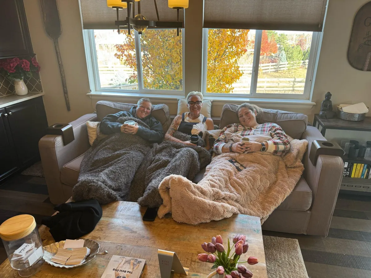 Three people relaxing together on a couch under cozy blankets, with drinks on the table and autumn leaves visible through the window behind them.