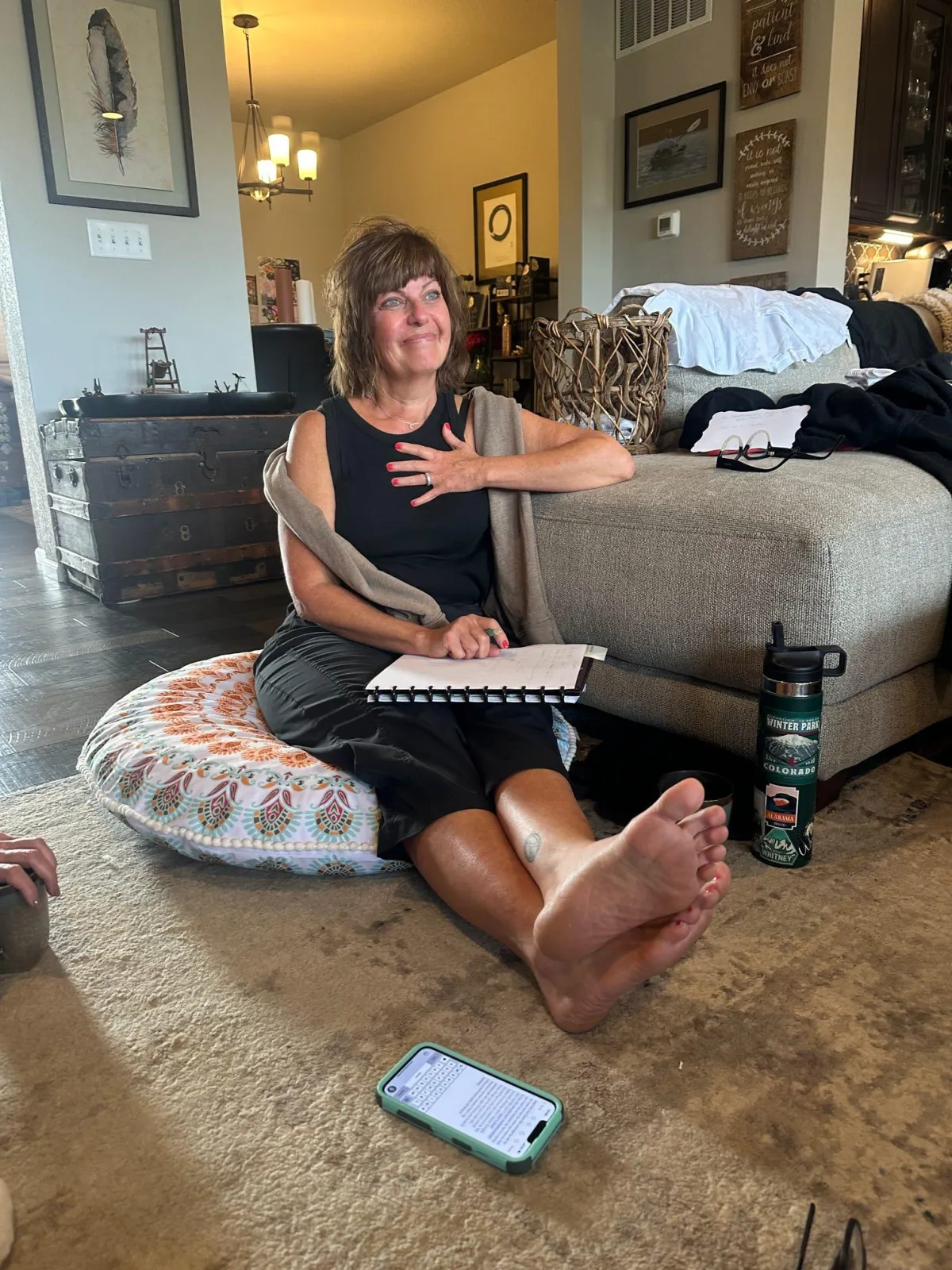 Woman sitting on a cushion on the floor in a living room, gesturing while speaking, with a notebook and water bottle nearby.
