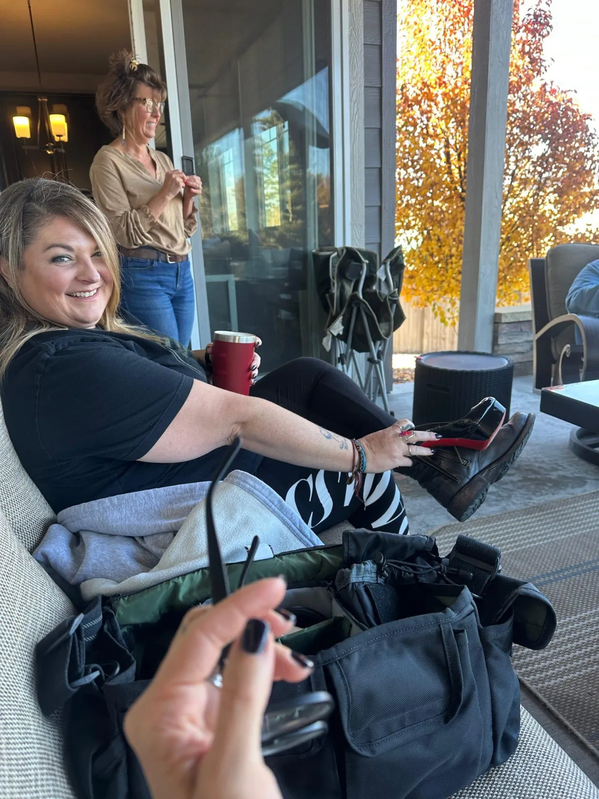 Woman smiling while sitting on a couch with bags and a drink nearby, as another woman stands by a large window with autumn leaves visible outside.