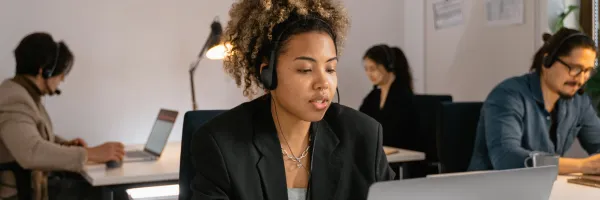 Woman in business attire on headset with three other people in the back ground.