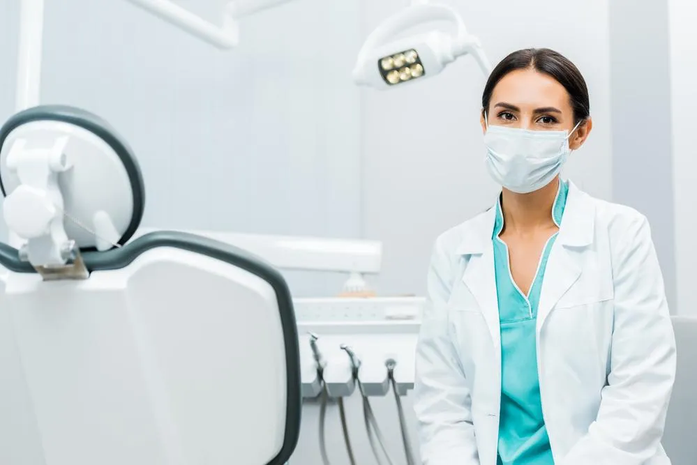 a masked female dental professional in a white lab coat and aqua colored scrub top seated next to the back of a dental chair
