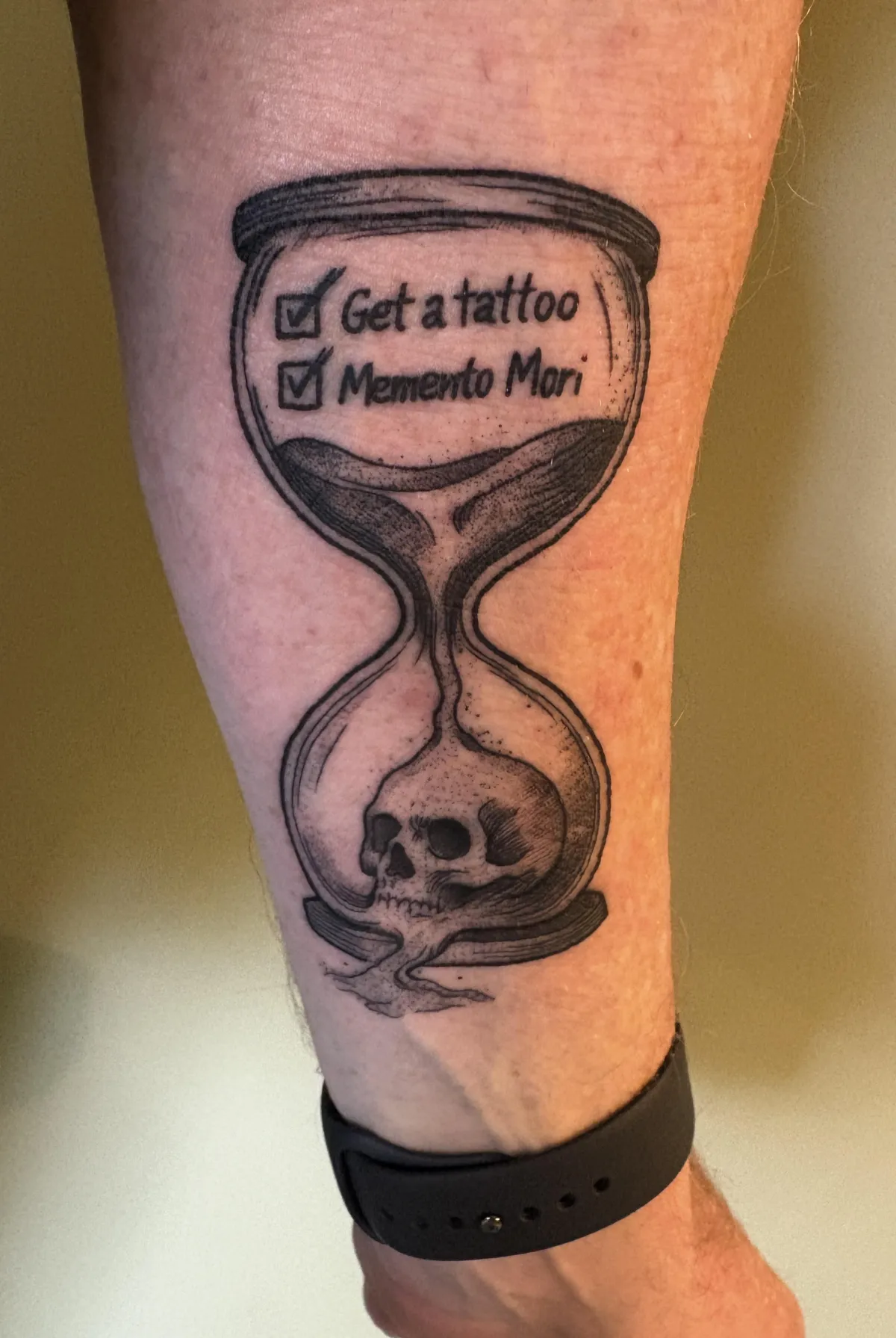 Black and gray hourglass tattoo with skull at bottom and two checked boxes at top reading "Get a tattoo" and "Memento Mori" - first tattoo at age 51