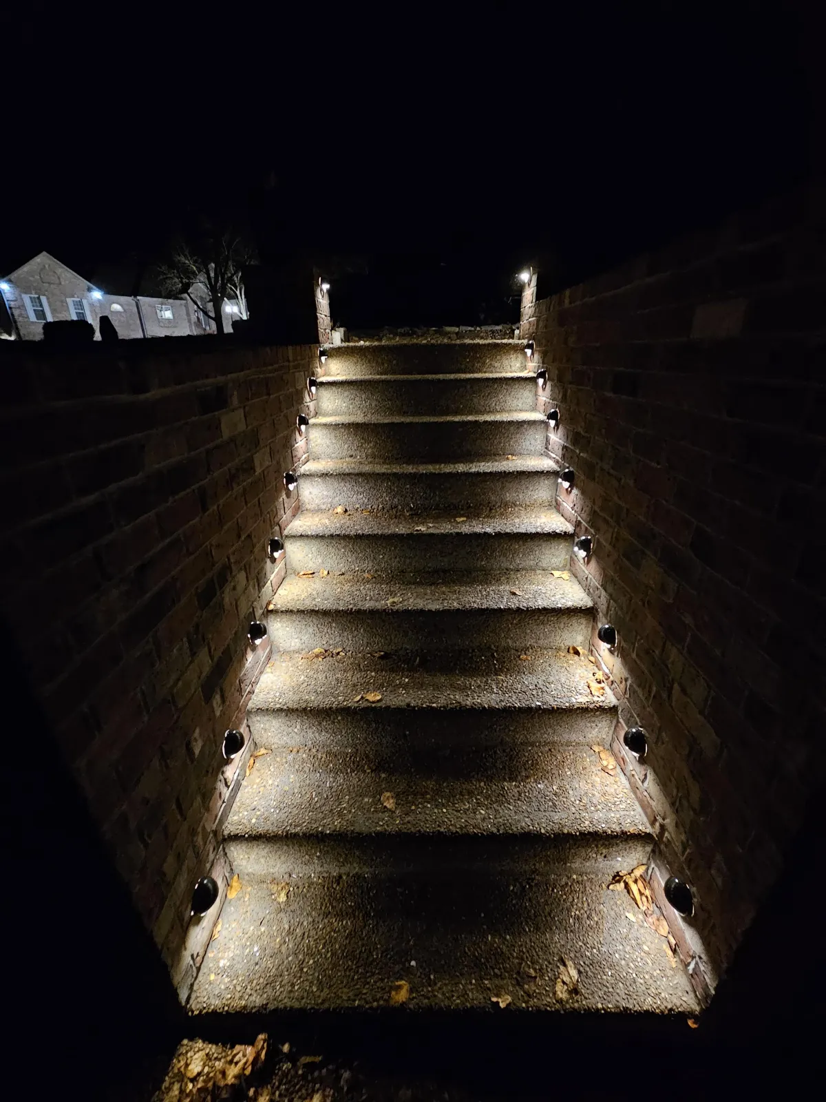 A Softly lit exposed aggregate stair well with brick siding. Each Step is lit on both sides with all brass circular hardscaping lights