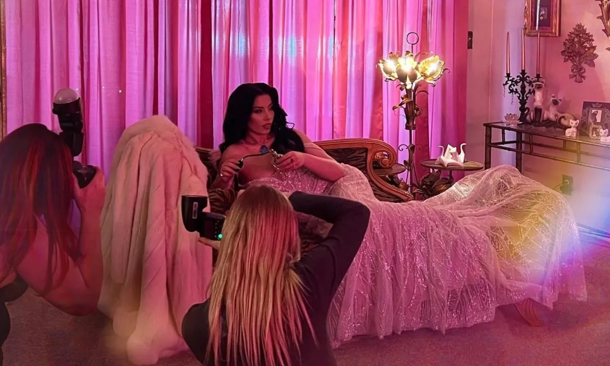 Royilda Medina lounges elegantly on a vintage-style couch in a softly lit pink room during a photoshoot, surrounded by two photographers capturing her in a glamorous gown with glowing backlight and warm tones.