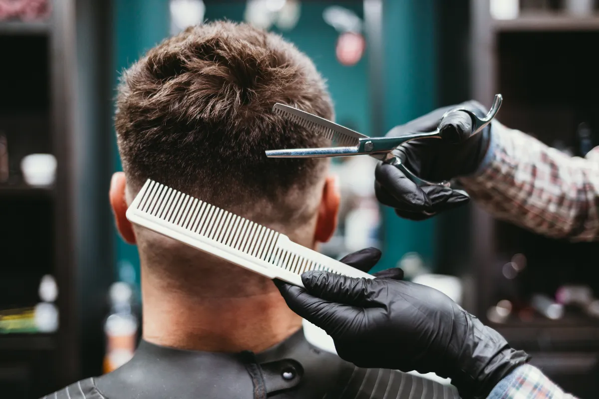 Barber giving a men’s haircut with scissors and comb, viewed from behind, inside a modern barbershop.