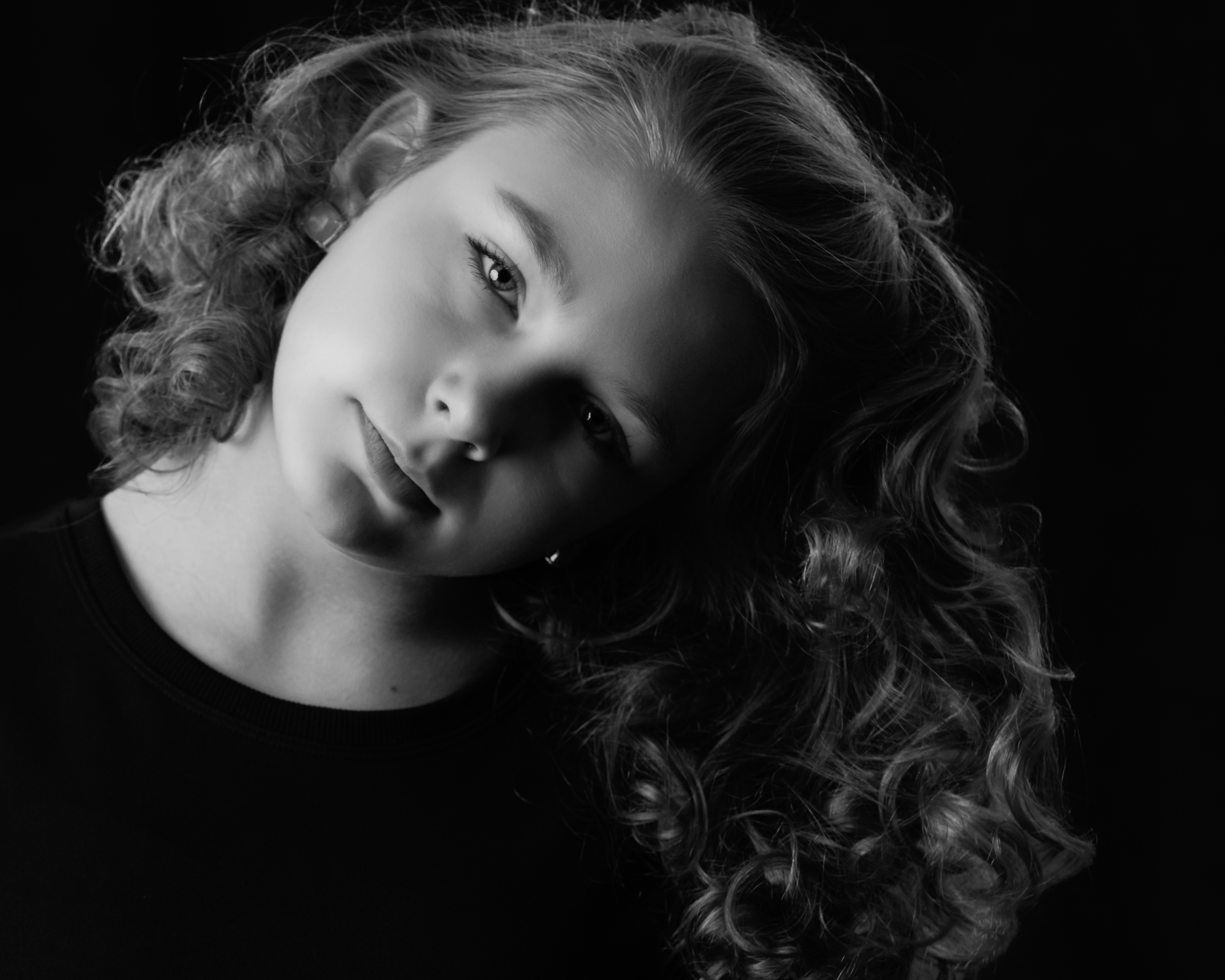 A black-and-white portrait of a young girl with softly curled hair, her head gently tilted as she gazes toward the camera against a dark background (Evidence), expressing a sense of calm curiosity and quiet openness (Emotion), reflecting the tenderness and self-awareness that emerge as she grows into her own sense of being (Elevation).