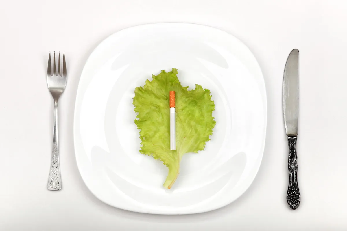 Un cigarrillo sobre una hoja de lechuga en un plato blanco, con cubiertos a ambos lados, simbolizando la relación entre dejar de fumar y la dieta