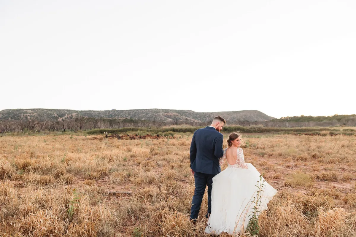 Bride and groom walking in a field at Sabrina Cedars in Abilene Texas with hills in the background, photographed by Double Knot Photography.
