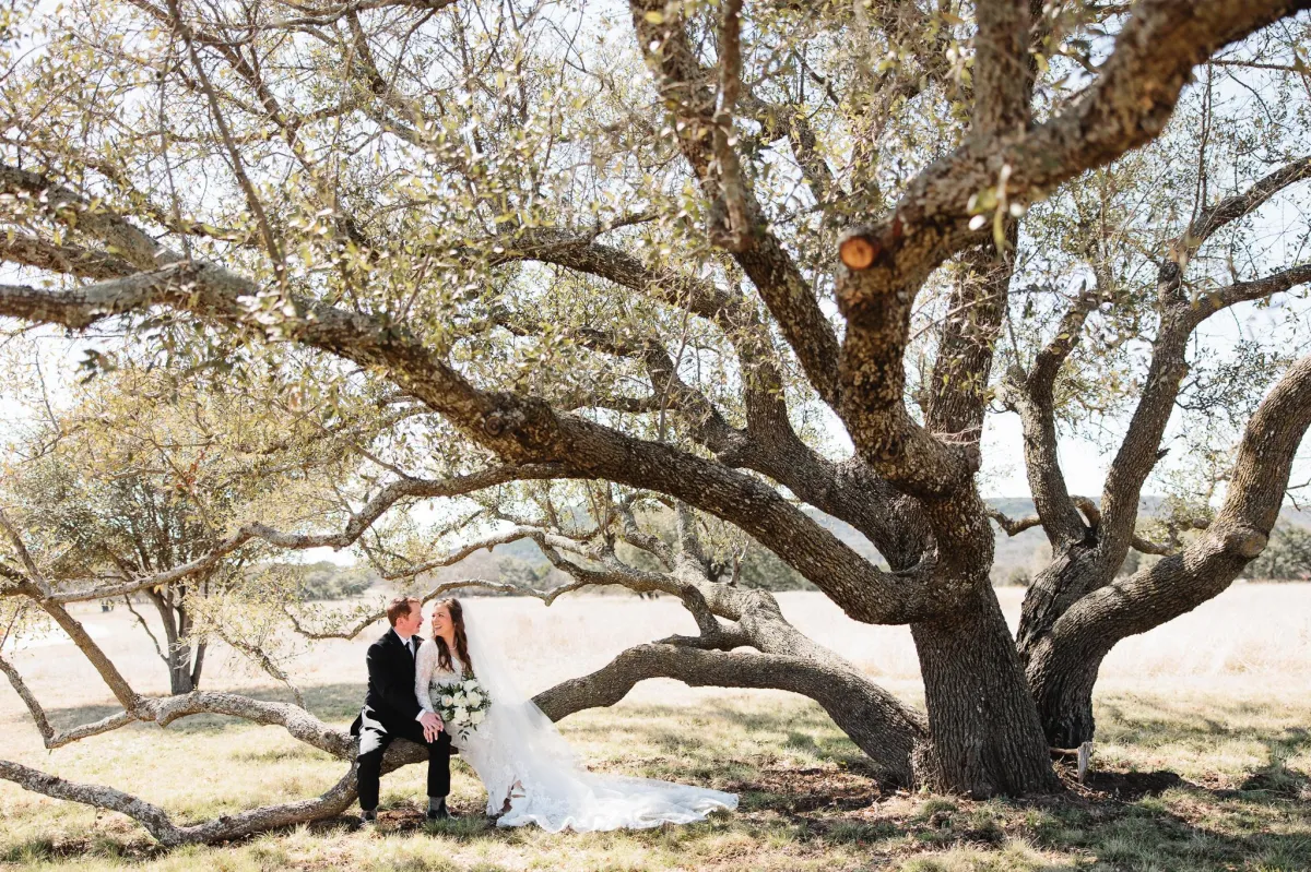 Bride and groom sitting under an oak tree with a bouquet, photographed by Double Knot Photography, showcasing an intimate wedding moment at Sparrow Creek Ranch in Graham Texas